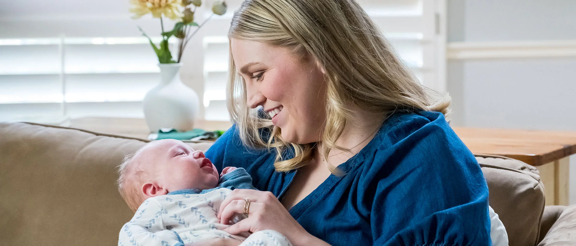 A smiling woman with blonde hair wearing a blue top sits on a couch, cradling a newborn baby in her arms. The baby is wearing a patterned white onesie.