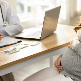 A pregnant person sits across from a doctor at a desk with ultrasound images, a notebook, and a laptop during a medical consultation.