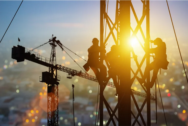 Silhouetted construction workers climb a tall metal structure at sunset, with cranes and blurred city lights in the background.
